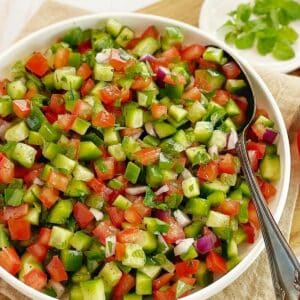 chopped tomato and cucumber salad in a white bowl with a spoon on a wooden board