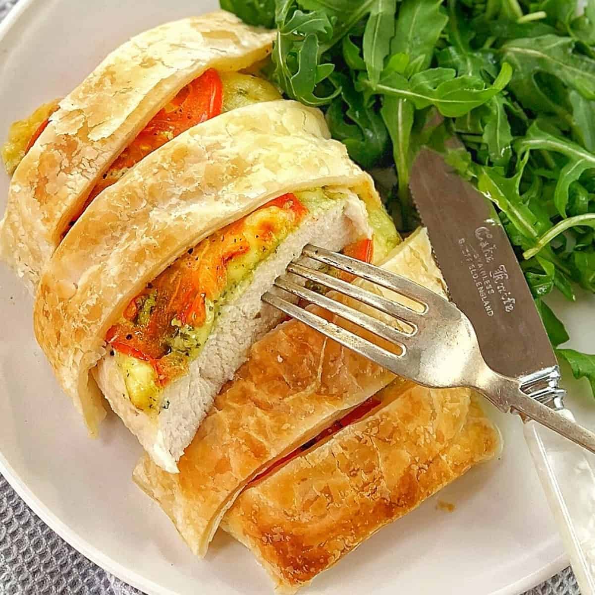 chicken en croute topped with pastry strips, mozzarella cheese and tomato on a white plate with arugula and a knife and fork