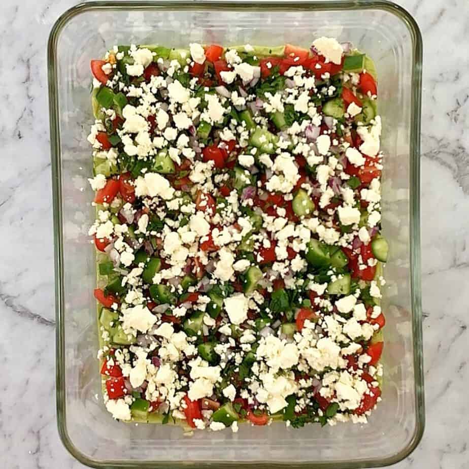 feta, parsley, cucumber, tomato on top of mashed avocado layer in a glass dish