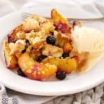 yellow peaches and blueberries with golden crumble topping on a white plate