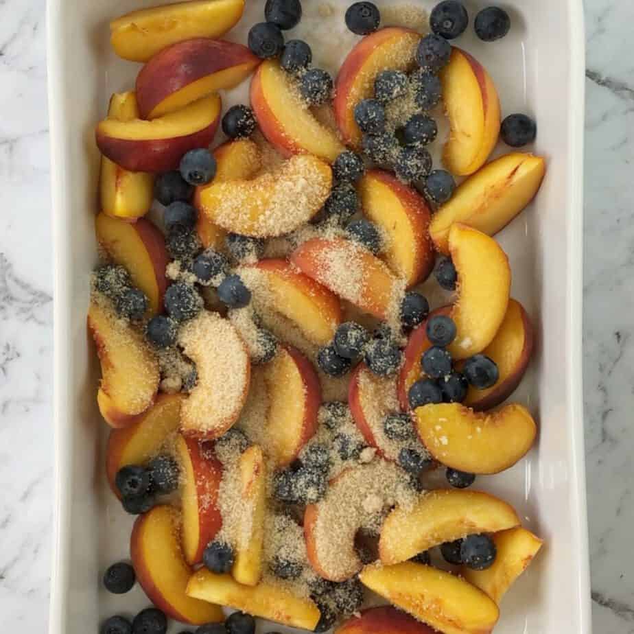 Peach slices and blueberries with sugar on top in a white baking dish