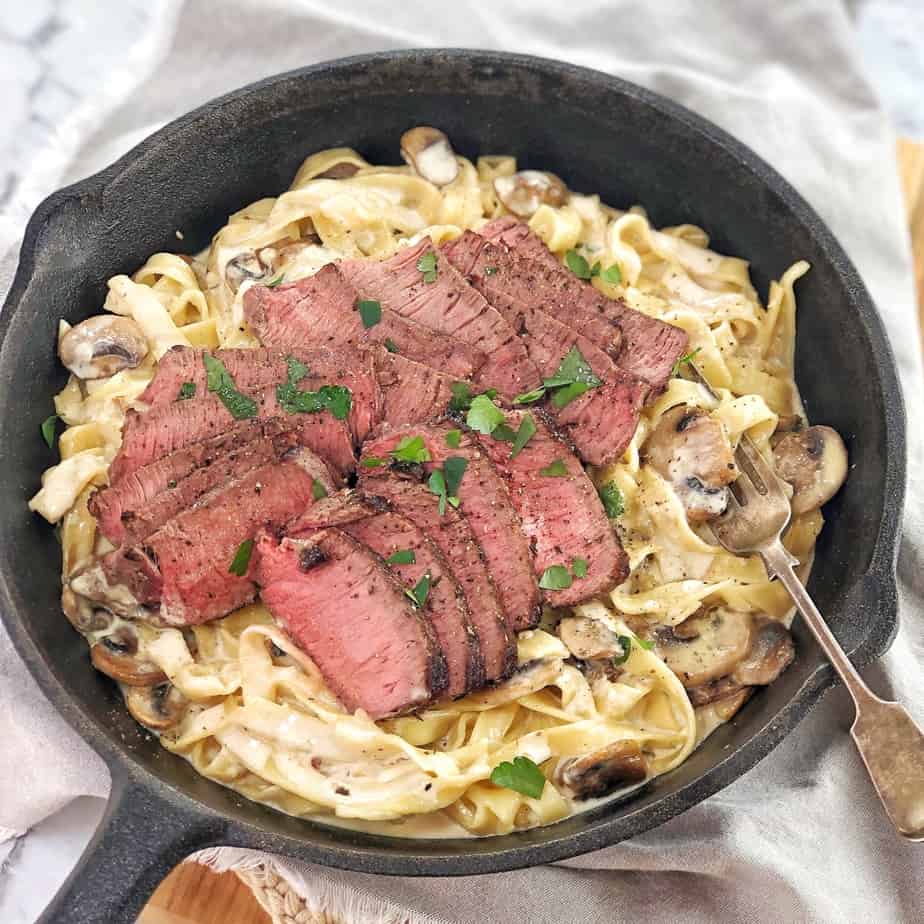 sliced steak sitting on pasta with a creamy sauce in black pan
