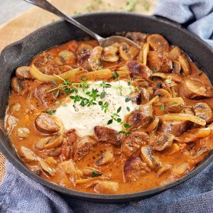 chopped sausages mushrooms and onions in a tomato sauce with sour cream on top in a black pan