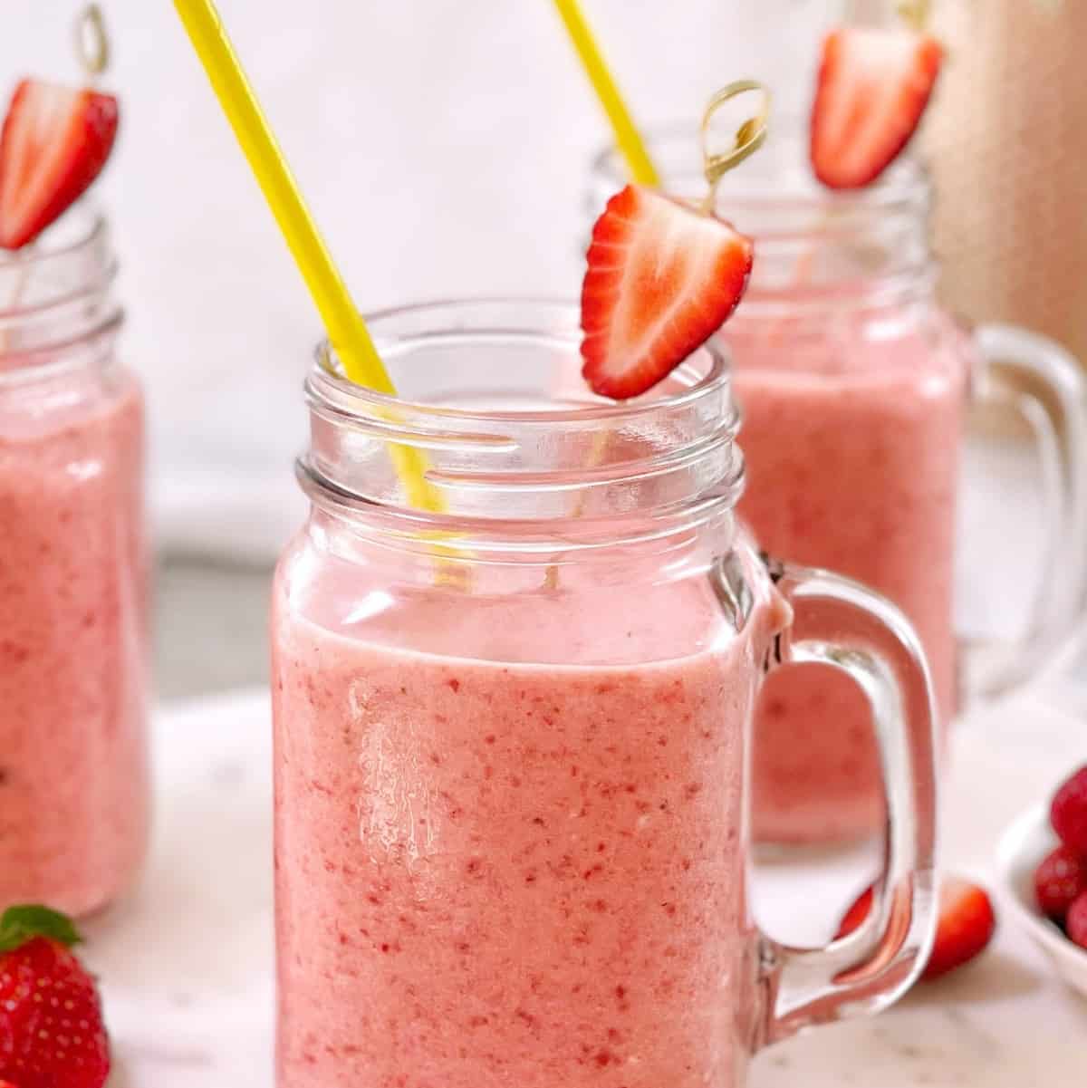 pink colored strawberry colada drinks in clear glasses decorated with strawberries and yellow straws on a marble board