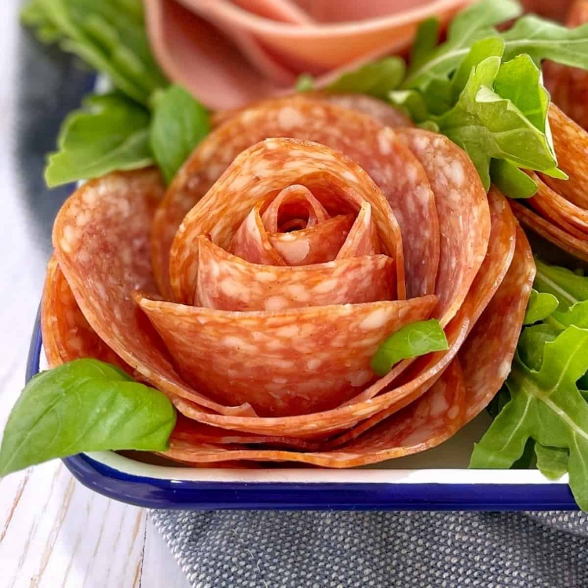 Pepperoni folded into the shape of a rose in a white tray with basil leaves and arugula.