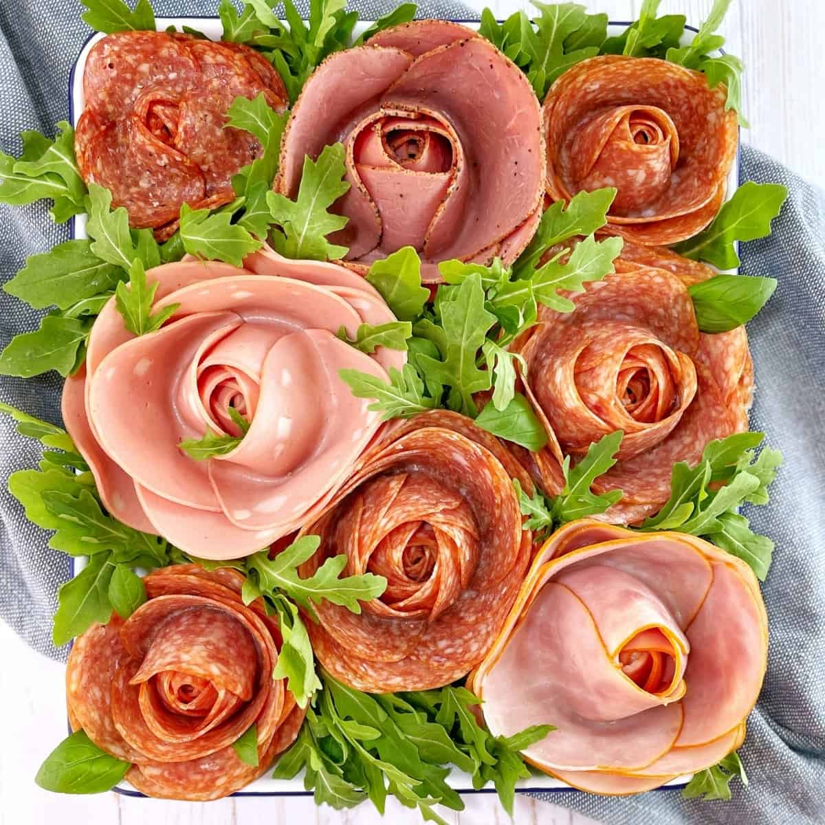 Several examples of meat roses made of pepperoni and other deli meats on a white tray with arugula.