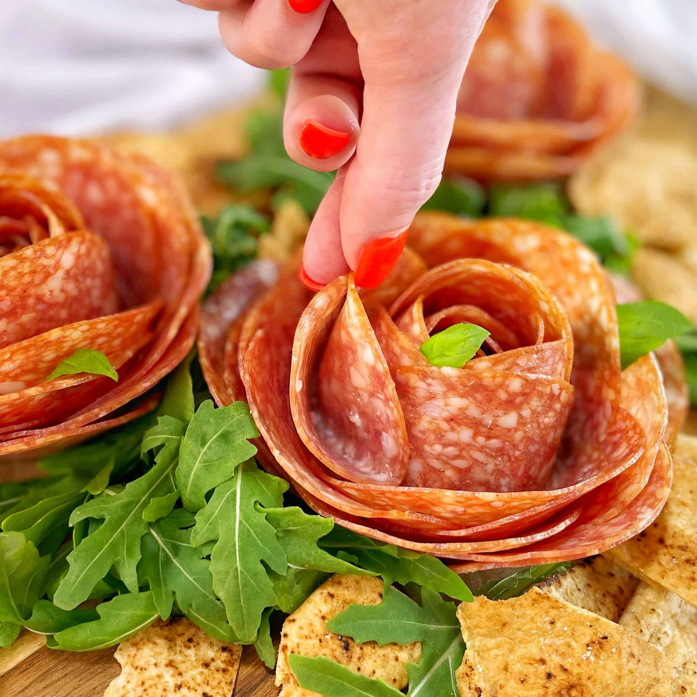 Fingers grasping a piece of pepperoni in a pepperoni rose on a wooden board with pita chips and arugula.