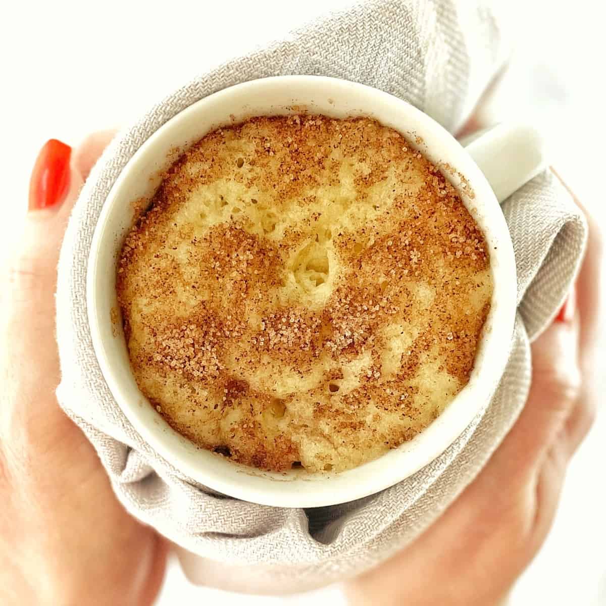 top down photograph of snickerdoodle cake in a white mug being held by two hands with a tea towel.