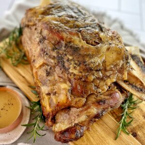 roast lamb shoulder on a wooden chopping board with rosemary and a small saucepan of gravy.