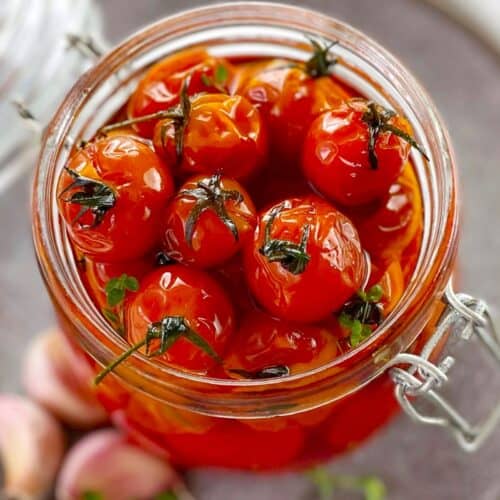 cooked cherry tomatoes in a glass jar on a gray plate with garlic cloves.