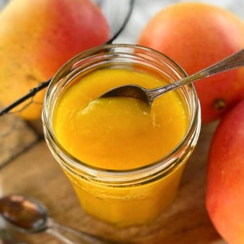 mango coulis sauce in a glass jar on a wooden board with fresh mangoes and a spoon.