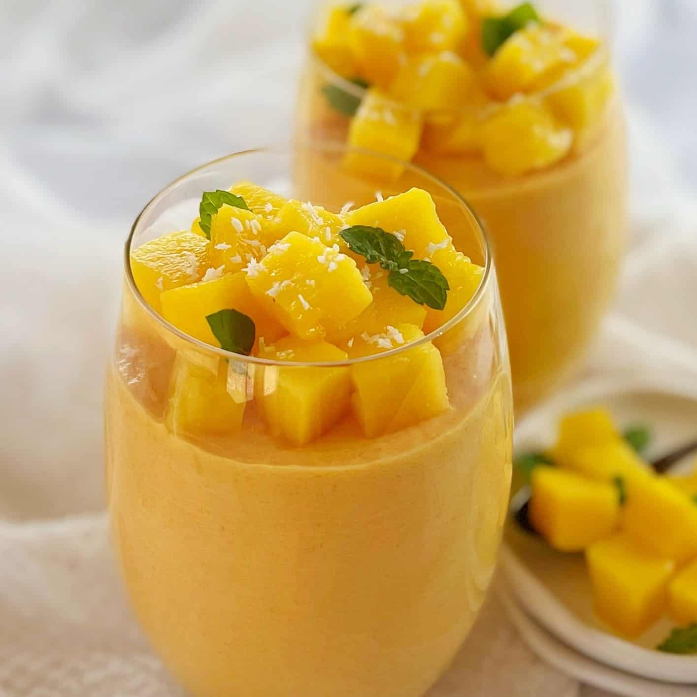 glasses filled with orange coloured mango mousse chopped mango on top with mint leaves and coconut.