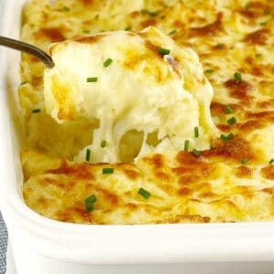 a spoon digging into baked mashed potato covered in melted cheese in a white casserole dish.