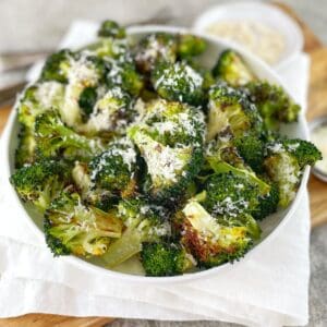 A white bowl with pieces of charred broccoli sprinkled with parmesan cheese on a wooden board with a white tea towel.