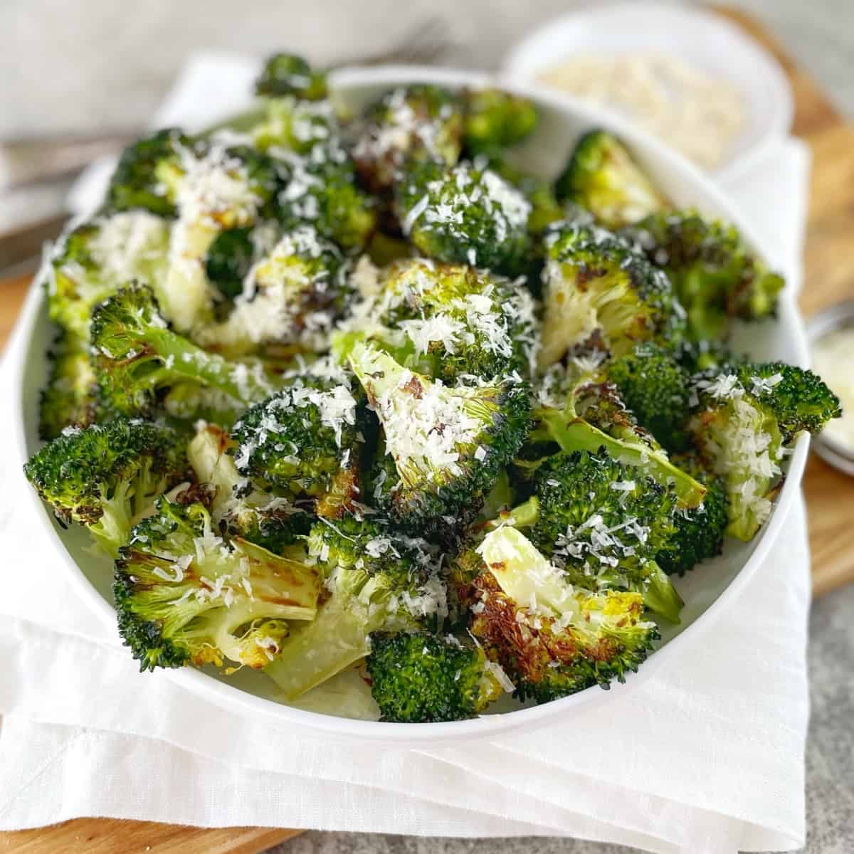 A white bowl with pieces of charred broccoli sprinkled with parmesan cheese on a wooden board with a