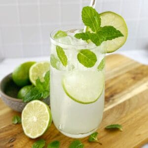 Cocktail in a tall clear glass garnished with lime and mint leaves on a wooden board.