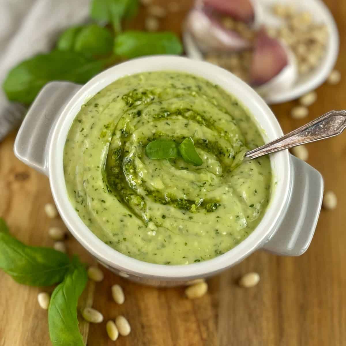 Mixture of mayonnaise and basil pesto in a white bowl on a wooden chopping board with a spoon.