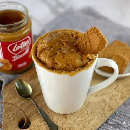 Cake made in a mug decorated with a cookie with a spoon and a jar of biscoff on a wooden board.