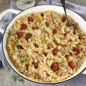 Risotto with chicken and chorizo in a white pan with a sprig of thyme and a spoon.