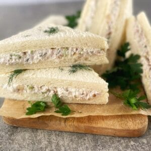 Triangle shaped chicken sandwiches on white bread sitting on a wooden board scattered with parsley and dill.