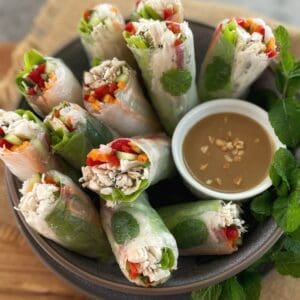 Rice paper rolls with chicken in a gray bowl with a brown dipping sauce and fresh mint on a wooden board.