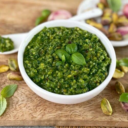 Green pesto in a white bowl on a wooden board with basil leaves and pistachios.