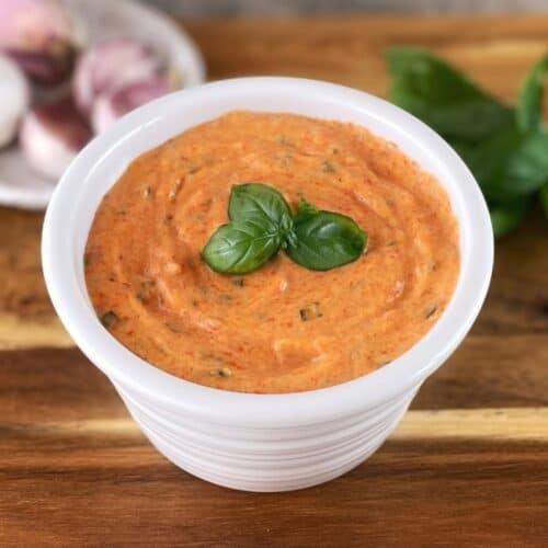 Creamy red pepper sauce in a white bowl on a wooden board with basil on top.