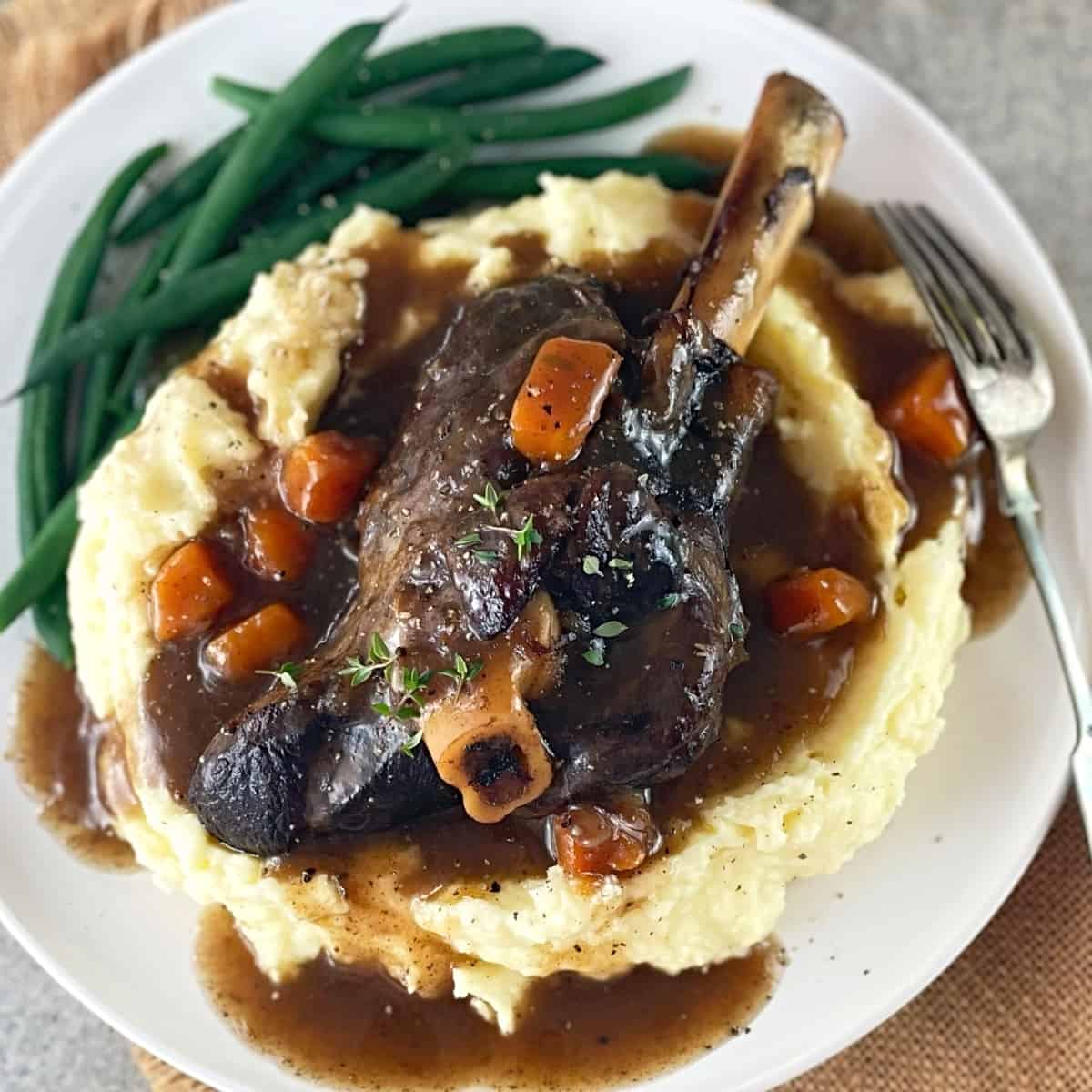 Lamb shank sitting on mashed potato on a white plate with green beans and a fork.