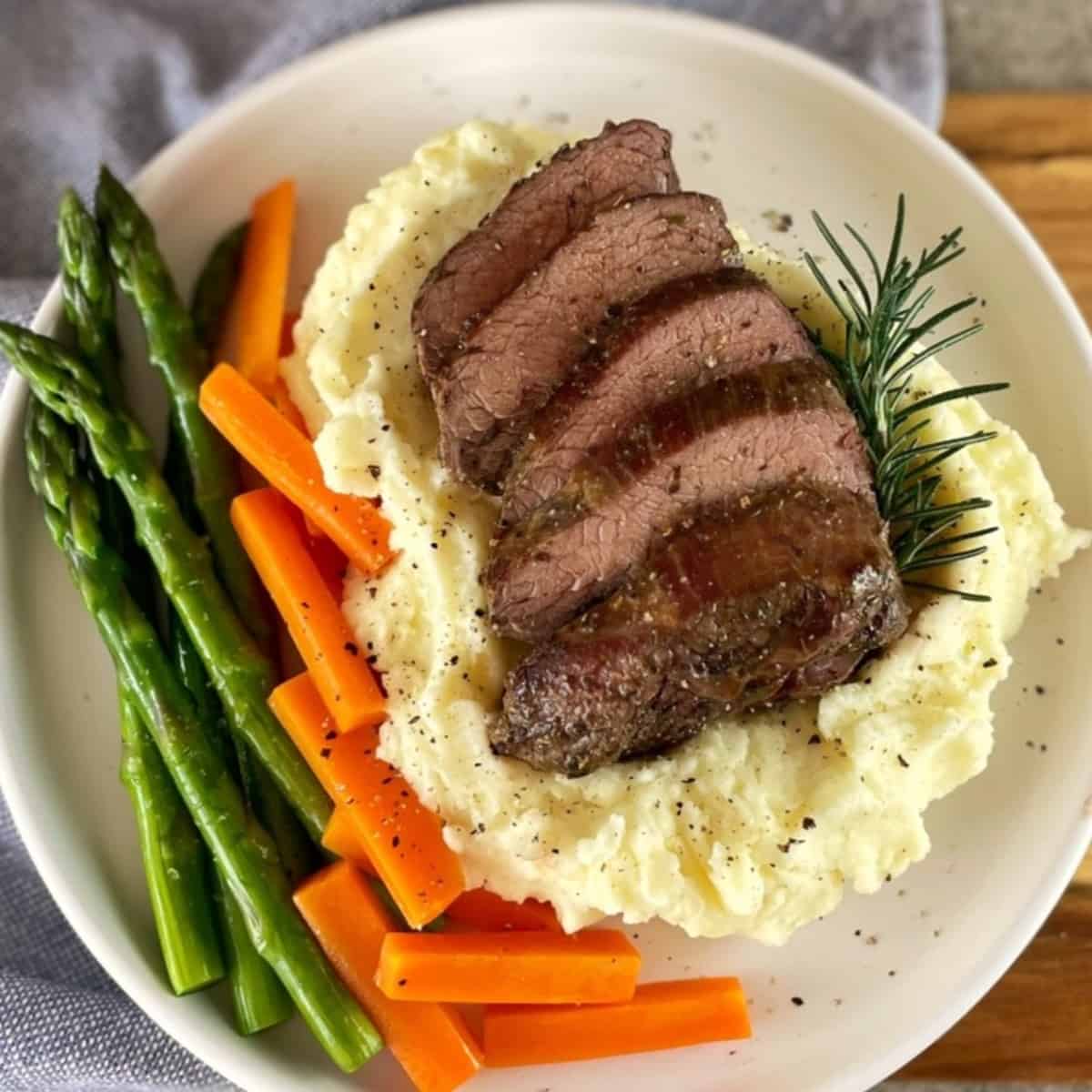 Sliced roast lamb rump sitting on mashed potatoes with asparagus and carrots on the side of the plat