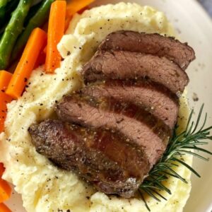 Sliced roast lamb rump sitting on mashed potatoes with asparagus and carrots on the side of the plate with a sprig of rosemary.