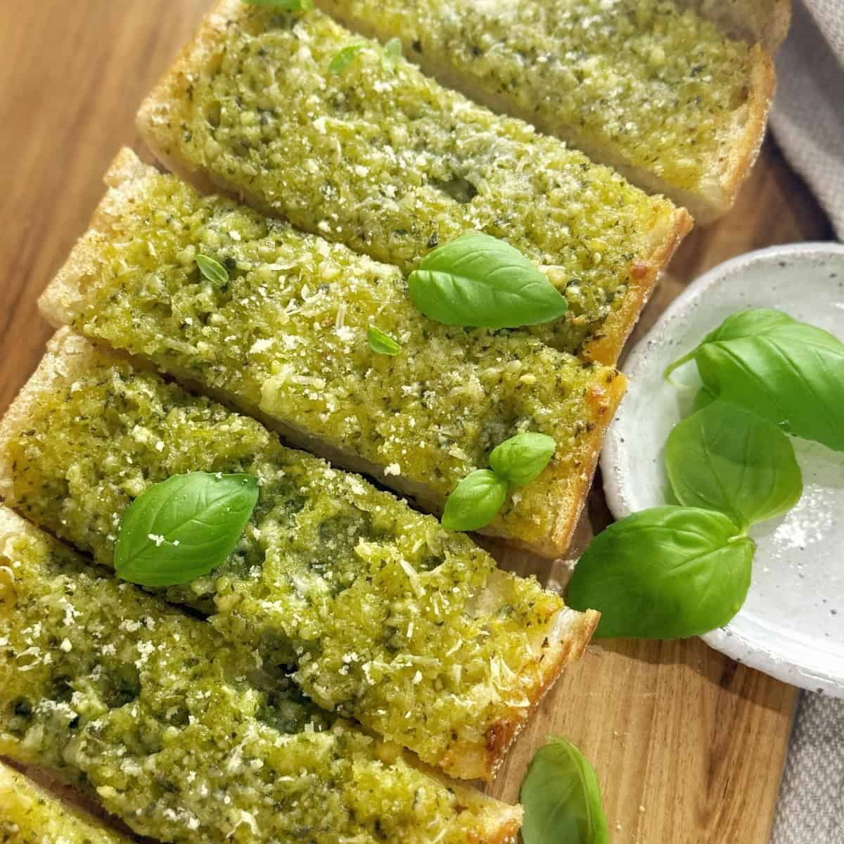 Garlic bread made with basil pesto sitting on a wooden board and garnished with basil leaves.